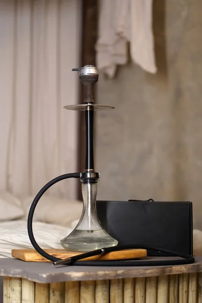 A large, luxurious metallic black hookah with a brown clay bowl with clear water and kaloud stands on a wooden gray table near the black box at home interior against the bed. The concept of rest.