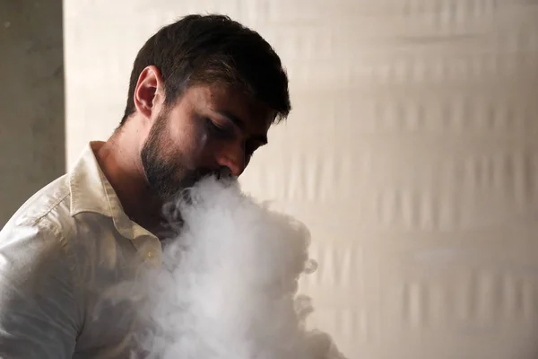 Portrait of a brutal bearded Caucasian man with short brown hair in a white shirt on blurred background. Handsome male smokes a hookah and exhales a cloud of smoke from his mouth with pleasure. Stock Picture