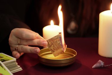 Cadı, yüzüklerle siyah pelerinli falcı. Büyü okuyup yakıyor. Tarot kartları, ametist taşları, karanlık ve gizemli arka planda beyaz mumlar. Okült, esrarengiz, kehanet ve cadı kavramı.