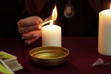Cadı, siyah mantolu falcı sihirli bir ayin ve yanık büyüsü yapıyor. Tarot kartları, ametist taşı, karanlık ve gizemli arka planda beyaz mumlar. Okült, esrarengiz, kehanet ve cadı kavramı.