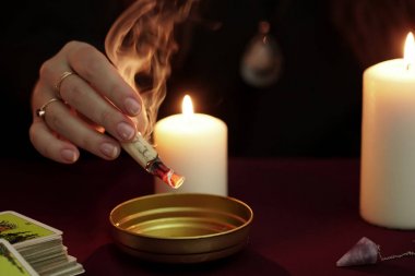 Cadı, siyah mantolu falcı elinde kasenin üzerinde büyüyle yanan bir kağıt tutuyor. Tarot kartları, ametist taşı, karanlık ve gizemli arka planda beyaz mumlar. Okült, esrarengiz, kehanet ve cadı kavramı