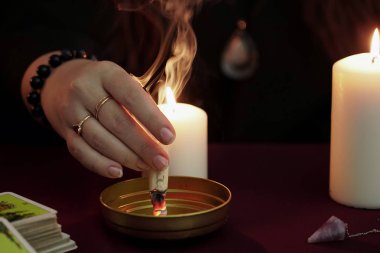 Cadı, siyah mantolu bir falcı ayin yapar ve kağıtları büyüyle yakar. Tarot kartları, ametist taşı, karanlık ve gizemli arka planda beyaz mumlar. Okült, esrarengiz, kehanet ve cadı kavramı.