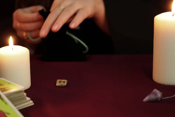 Cadı siyah mantolu bir falcı çantadan antik rünleri çıkartıyor. Tarot kartları, ametist taşları, karanlık ve gizemli arka planda beyaz mumlar. Okült, esrarengiz, kehanet ve cadı kavramı.