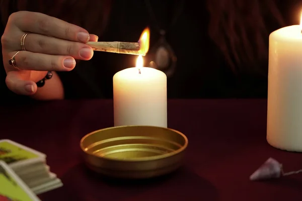 La bruja es adivina en manto negro con anillos evoca papel quemado con un hechizo. Tarjetas del tarot, piedras de amatista, velas blancas sobre fondo oscuro. Concepto oculto, esotérico, adivinación y wicca . — Foto de Stock