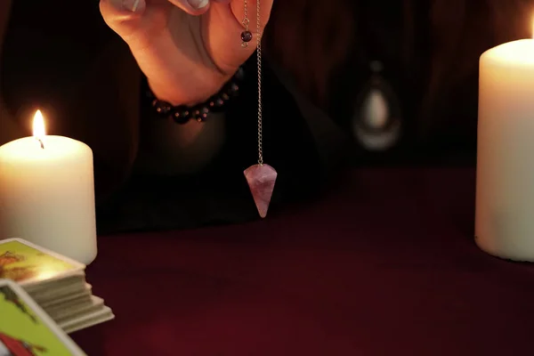 La bruja es adivina en túnica negra sostiene en la mano péndulo de amatista en la cadena. Tarjetas del tarot, piedra amatista, velas blancas sobre fondo místico oscuro. Concepto oculto, esotérico, adivinación y wicca . — Foto de Stock