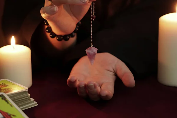 Cadı, siyah cüppeli falcı elinde ametist sarkacı tutuyor. Tarot kartları, ametist taşı, karanlık ve gizemli arka planda beyaz mumlar. Okült, esrarengiz, kehanet ve cadı kavramı.
