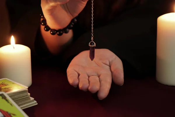 Heksa er spåkone i svart kappe holder ametyst-pendel på kjetting over hånd. Tarotkort, ametyststein, hvite lys på mørk, mystisk bakgrunn. Okkulte, esoteriske, spådommer og wicca-konseptet . – stockfoto