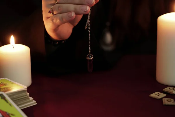 La bruja con un manto negro sostiene en su mano un péndulo de amatista de piedra en una cadena. Tarjetas del tarot, runas antiguas y velas blancas. Concepto oculto, esotérico, adivino y Wicca. Fondo místico . — Foto de Stock