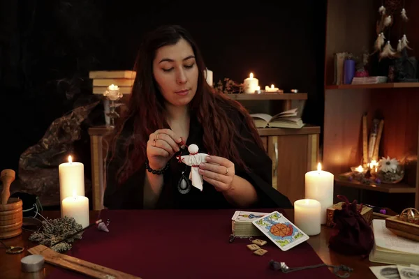 Una bruja es adivina en manto negro sostiene muñeco vudú con hilos rojos. Ritual mágico. Tarjetas del tarot, velas blancas en la mesa. Oculta, esotérica, adivinación y concepto Wicca. Fondo místico . — Foto de Stock