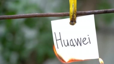 Bulanık arka planda sarı clothespin üzerinde beyaz etiket üzerinde Yazıt. Kelime Huawei metal çubuk asılı kağıt. Mans el kırmızı çakmak sağdaki kelime tutuşturuyor kullanın. Kağıt yanık.