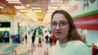 Young blond man with acne on his face in a purple pullover kisses beautiful girl in round glasses in white woolen sweater and smiles against blurred background of people walking in the store.