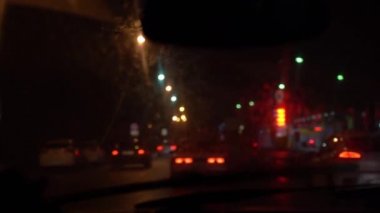 Defokus arabanın içinden kirli ıslak bir ön cam dan dışa görünüm. Araba gece yolunda bir araba akışı içinde hareket eder. Ön cam silecekleri camı yıkar. Işık ve araba yansımaları.