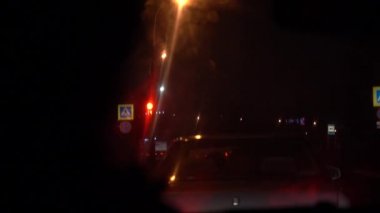 Arabanın içinden kirli ıslak bir ön camdan dışa doğru görünüm. Araba gece yolundaki trafik ışıklarında durur. Saniyeler 15'ten 8'e kadar sayılır. Işık ve araba yansımaları.