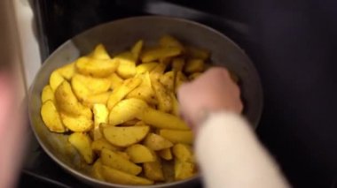 Siyah plastik pişirme çatal kullanarak beyaz pullover bir kadın el evde fırında fırın tepsisine ızgara sarı sos baharat ile kızarmış patates lezzetli dilimleri karıştırır. Duman buharlaşır..