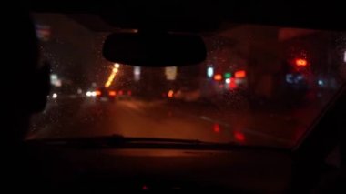 Isliç bir ön camın iç görünümü. Araba gece yoluna çıkıyor. Sürücü sağa, sola döner ve eliyle sollama hareketi yapar. Cama odaklan. Araba farlarının bulanık yansımaları.