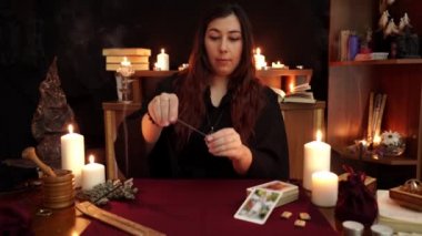 Bir cadı cüppe falcı kara büyü bir ayin gerçekleştirir. Sarkaç evet diyor. Büyülü bir ritüel. Tarot kartları, beyaz mumlar ve rünler. Gizli, ezoterik ve kehanet kavramı. Mistik arka plan.