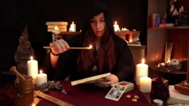 Bir cadı siyah mantolu falcı, kâğıt üzerine mumdan büyü ve damla balmumu okur. Büyülü bir ritüel. Tarot kartları, mumlar ve koyu arka planda rünler. Gizli, ezoterik ve kehanet kavramı.