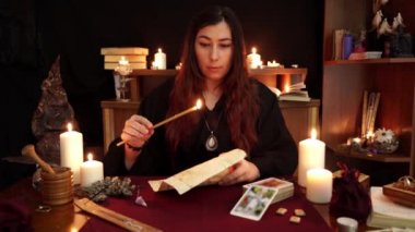 Bir cadı siyah mantolu falcı kilise mum tutuşturmak, büyü okur ve büyü ile kağıt üzerine balmumu damla. Tarot kartları, mumlar ve koyu arka planda rünler. Gizli, ezoterik ve kehanet kavramı