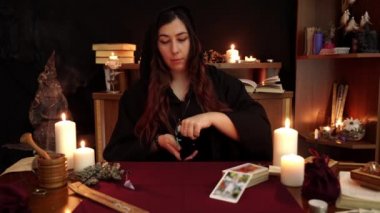 Cadı, elinde yüzük olan siyah mantolu bir falcı. Masaya antik mühürler koyuyor. Tarot kartları, ametist taşları, karanlık ve gizemli arka planda beyaz mumlar. Okült, esrarengiz, kehanet ve cadı kavramı.