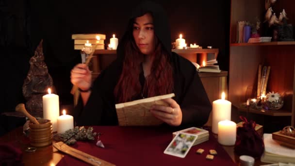 魔女は黒いローブの占い師で教会のろうそくに火をつけ 呪文で紙に呪文とドリップワックスを読みます 魔法の儀式タロットカード キャンドル ルーンをテーブルの上に置きます オカルトと占いの概念 — ストック動画