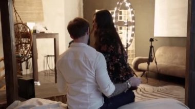 Bir ipek cüppe seksi bir kız bir adamın diz oturur ve onu uzun siyah saç düzer. Beyaz buruşturulmuş gömlek sakallı Kafkas erkek evde yatak odasında bej ahşap yatakta kadın kucaklar. Geri görünüm.