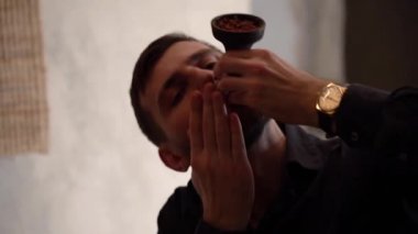 Siyah gömlekli yakışıklı beyaz sakallı bir adam, tütünün acısından dolayı tütün ve grimaces yakmak için kil bir kase üfler beyaz bir taş duvara. Nargile temizleme konsepti.