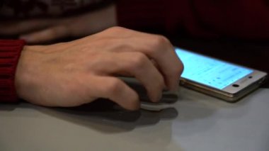 Kırmızı bir kazak ve dahil ekranlı beyaz bir telefon izleri ve damarları ile genç bir adamın elini yakın çekim. Adam gergin, parmaklarını masaya vuruyor ve indirmeyi bekliyor..