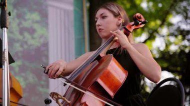 Siyah elbiseli çekici sarışın kadın çello klasik müzik solosu çalıyor. Güzel kız gün batımında açık havada sokak müzisyenleri topluluğunda gösteri yapıyor. Parkta senfoni orkestrası çalıyor.