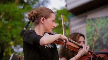 Açık havada sokak müzisyenleri topluluğu. Siyah elbiseli beyaz beyaz kadın gün batımında güzel ahşap keman klasik müzik çalmak. Senfoni orkestrası parkta ilk keman bölümü performans
