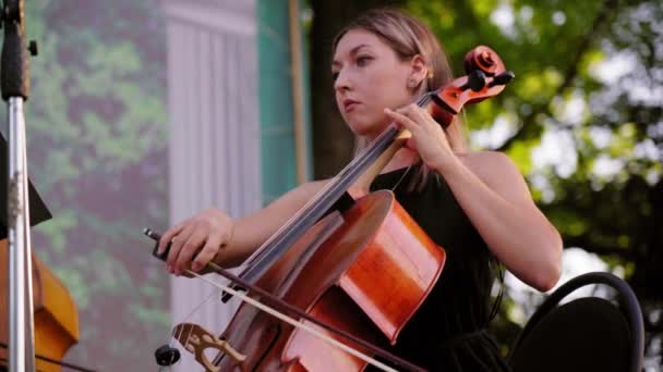 穿着黑色衣服的迷人金发女人演奏大提琴古典音乐独奏 日落时 美丽的姑娘在露天表演街头音乐家的合唱 在公园表演的交响乐团 — 图库视频影像