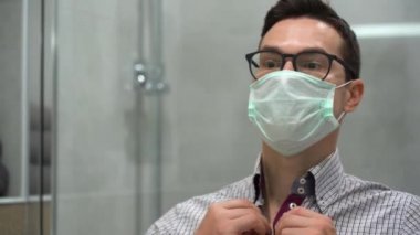 Genç, güzel, beyaz tenli, koruyucu tıbbi yüz maskesi takan, gri ekose gömlekli, güzel, esmer adam aynada kendine hayran. Erkekler tuvaletinde ofis çalışanının portresi.