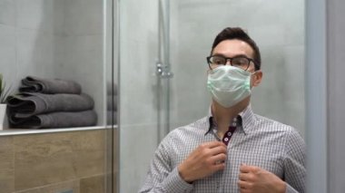 Tek kullanımlık turkuaz koruyucu maskeli, siyah gözlüklü, ekose gömlekli, beyaz, yakışıklı, esmer bir adam sabah gri banyonun arka planındaki aynaya bakıyor.