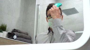 Genç, tatlı esmer adam klasik ekose gömlek, siyah gözlük, koruyucu sağlık maskesi aynaya bak, tarak, kısa kahverengi saçlarını arka plandaki gri tuvalette düzeltir. Salgın. İç görünüm.