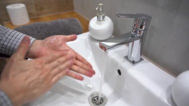 Gri tişörtlü, temiz elleri temiz metal musluk veya musluktan gelen saf sıcak suya sarılmış, parmaklarını ovuşturan erkek evde sık sık beyaz lavaboda sabunla yıkanıyor. Coronavirus salgını önleme. Sabah tuvaleti. İç görünüm.
