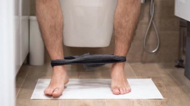 Gri iç çamaşırlı adam klozete oturur, beyaz paspasın üzerinde durur, kıllı bacaklarını sallar, banyoda el ele kağıt tutuşturur. İçeriden bak, kapat. Sabah rutini. Kahverengi fayansların arka planı. İç görünüm.