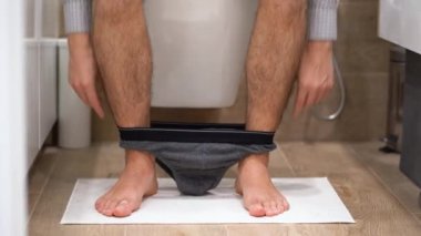 Ekose gömlekli genç bir işadamı beyaz bir klozetin üzerine oturur, gri iç çamaşırını çıkarır, pantolonunu giyer, ayağa kalkar ve gider ya da kahverengi banyoyu kapatır. Yüzü olmadan. İç görünüm.