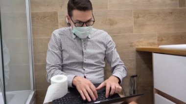 Gri gömlekli esmer iş adamı girişimci, dizüstü bilgisayar kullanarak koruyucu tıbbi yüz maskesi takıyor, tuvalette notlar yazıyor. Covid-19 karantinası sırasında bilgisayarla çalışan bir adam. İç görünüm.