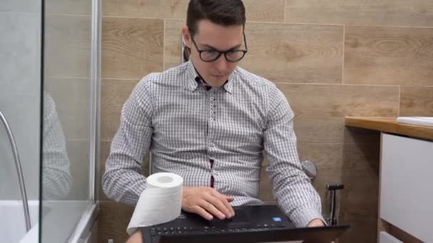 年轻英俊的白种人白种人办公室职员穿着格子花灰色衬衫 戴着黑色眼镜 坐在马桶上 用笔记本电脑冲浪 上网就近 一个带着电脑的男人在浴室里安静下来放松 内部视图 — 图库视频影像