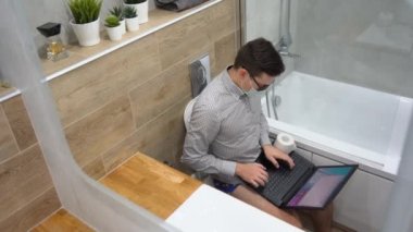 Genç esmer programcı iç çamaşırı, siyah gözlük, gri gömlek, koruyucu yüz maskesi takıyor uzaktan çalışıyor, dizüstü bilgisayar kullanıyor, yazı yazıyor, tuvaletin arka planında beyaz tuvalette oturuyor. Üst Manzara. İç görünüm.