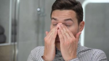 Ekose gömlekli yakışıklı beyaz adam aynaya bakıyor, kaşındıran kahverengi gözlerine masaj yapıyor, gri arka planda siyah gözlük takıyor. Yorgun ve stresli hissediyorum. Görme sorunu, görme sorunu. İç görünüm.