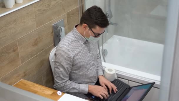 年轻的白人黑发企业家 戴着黑色眼镜 灰色格子花衬衫 面罩坐在白色马桶上 使用笔记本电脑 在厕所工作 隔离时间的远程家庭工作 内部视图 — 图库视频影像