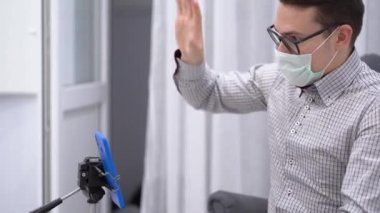 Genç esmer adam gri klasik ekose gömlek, siyah gözlük, tıbbi yüz maskesi giyer video görüşmesi için tripoda telefon takar. Erkek, patronla internette tanışıyor, iş buluyor. Uzaktan kumandalı iş. Kilitleyin. Yan görünüm.