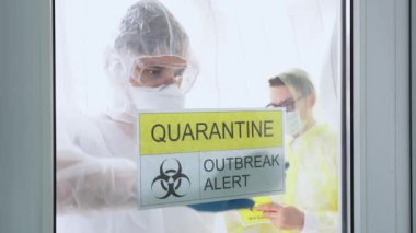 Erkek doktor yüz maskesi takıyor, güvenlik kıyafeti giyiyor, başlık dikkat dağıtıyor karantina, salgın alarmı. İnsanlar sosyal mesafeyi koruyorlar, hastanenin beyaz odasında dikiliyorlar. Koruma konsepti. İç görünüm.