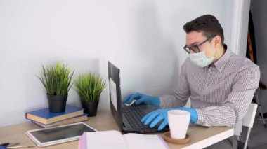 Ekose gömlekli, mavi eldivenli, koruyucu maske takan, siyah gözlüklü, dizüstü bilgisayarda çalışan genç bir çalışan. Adam kahve, telefon, kitap, not defteri ve tabletle masada oturuyor. İş yeri. İç görünüm.