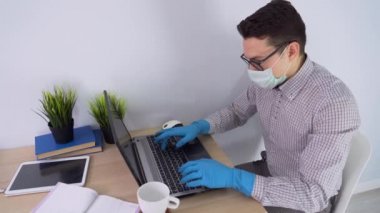 Siyah gözlüklü, koruyucu maskeli, lastik eldivenli, ekose gömlekli yakışıklı, beyaz tenli esmer adam masanın yanına oturur, dizüstü bilgisayar kullanır, yakın çekim yapar. Tecrit sırasında evden çalışmak. Üst görünüm.