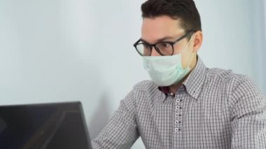 Beyaz tenli yakışıklı esmer adam siyah gözlük takıyor, tek kullanımlık tıbbi maske takıyor, ekose gömlek uzaktan çalışıyor, dizüstü bilgisayar kullanıyor, arka plandaki gri duvara yazı yazıyor. 