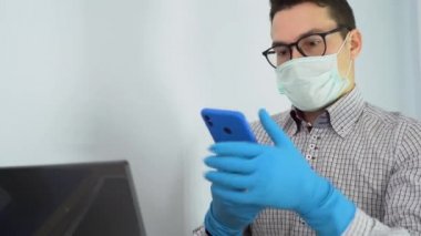 Turkuaz koruyucu tıbbi yüz maskeli, gri gömlekli, mavi antibakteriyel lastik eldivenli yakışıklı girişimci dizüstü bilgisayar kullanıyor, telefonda konuşuyor. Karantina zamanında çalış. Kilitle..