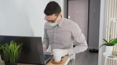 Adam gözlük takıyor, koruyucu sağlık maskesi takıyor, ellerinde beyaz lastik eldivenler var. Adam masadaki sandalyeye oturur, dizüstü bilgisayar kullanır, iş ortağıyla iletişim kurar. İşçi tablet ekranında grafiği göster