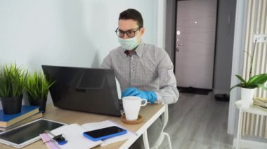 Genç beyaz iş adamı masada çalışıyor, dizüstü bilgisayar kullanıyor, maskesini çıkarıyor, beyaz bardak alıyor, ev mobilyalarında kahve içiyor. Adam klasik gömlek, siyah gözlük, mavi koruyucu eldiven giyiyor..