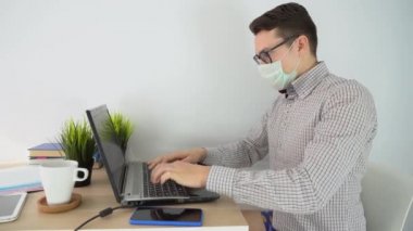 Kahverengi kısa saçlı esmer adam siyah gözlük, mavi şort, eğlenceli yeşil çorap, gri iş gömleği, koruyucu tek kullanımlık tıbbi maske, dizüstü bilgisayar, yazı, internet sörfü